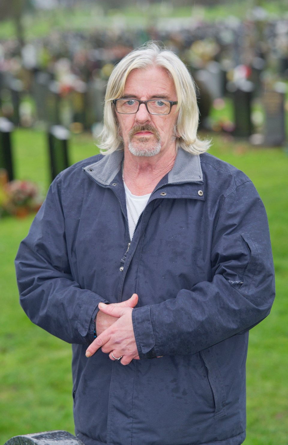 a man wearing glasses and a blue jacket stands in a cemetery