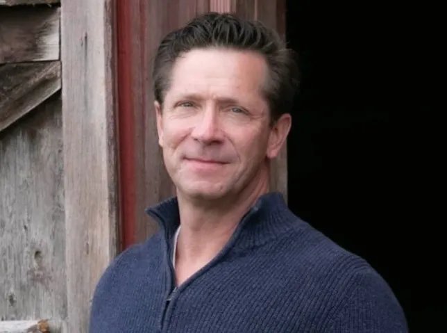 a man in a blue sweater is standing in front of a wooden building .