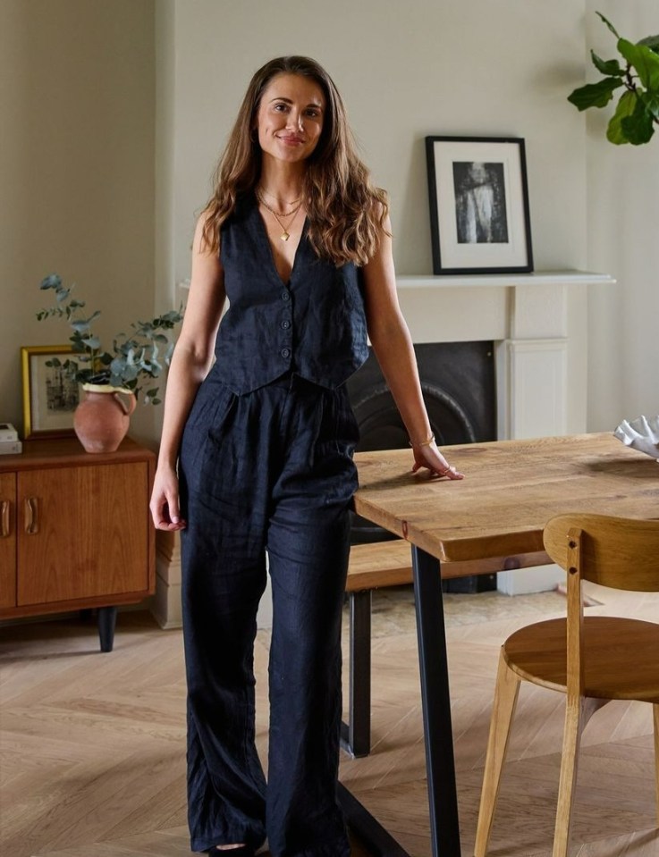 a woman in a black jumpsuit stands in front of a wooden table