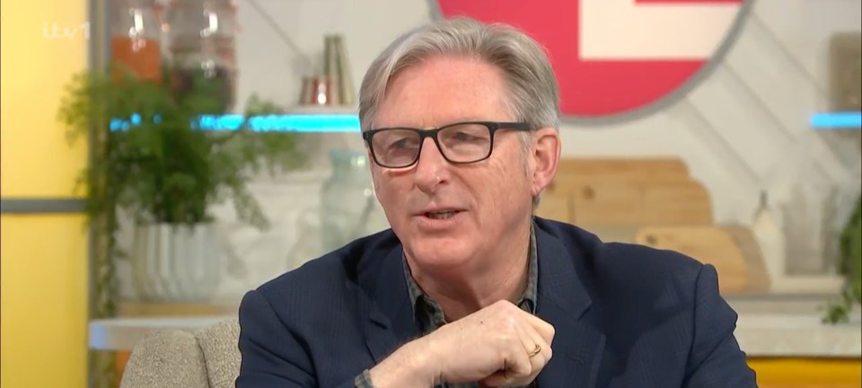 a man wearing glasses and a blue suit is sitting in front of a red sign that says itv