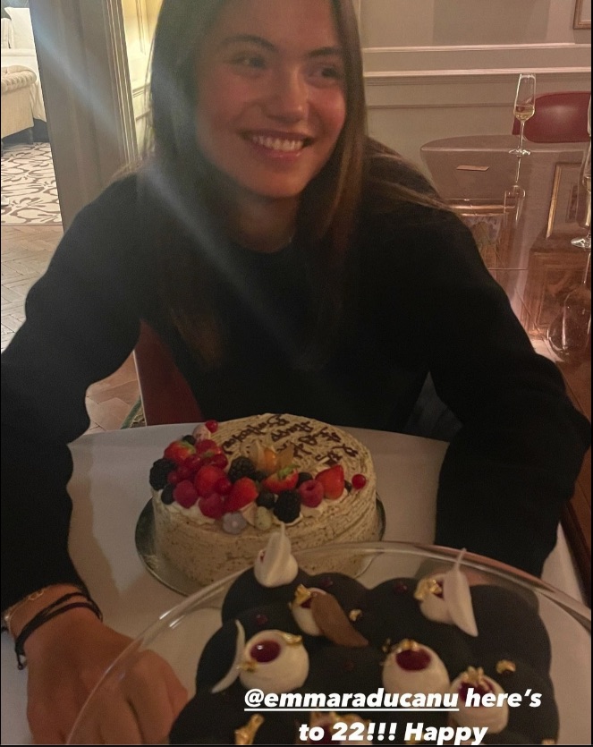 a woman sitting at a table with a cake and a caption that says emmaraducanu here 's to 22 !!! happy