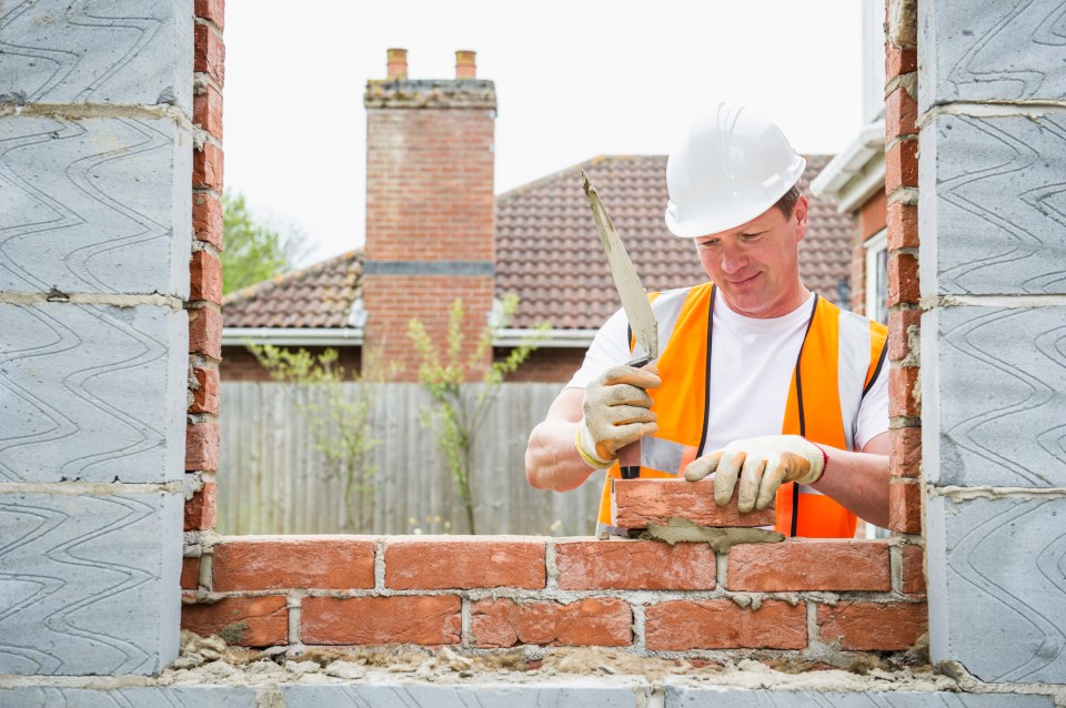 a man wearing a hard hat and orange vest is working on a brick wall