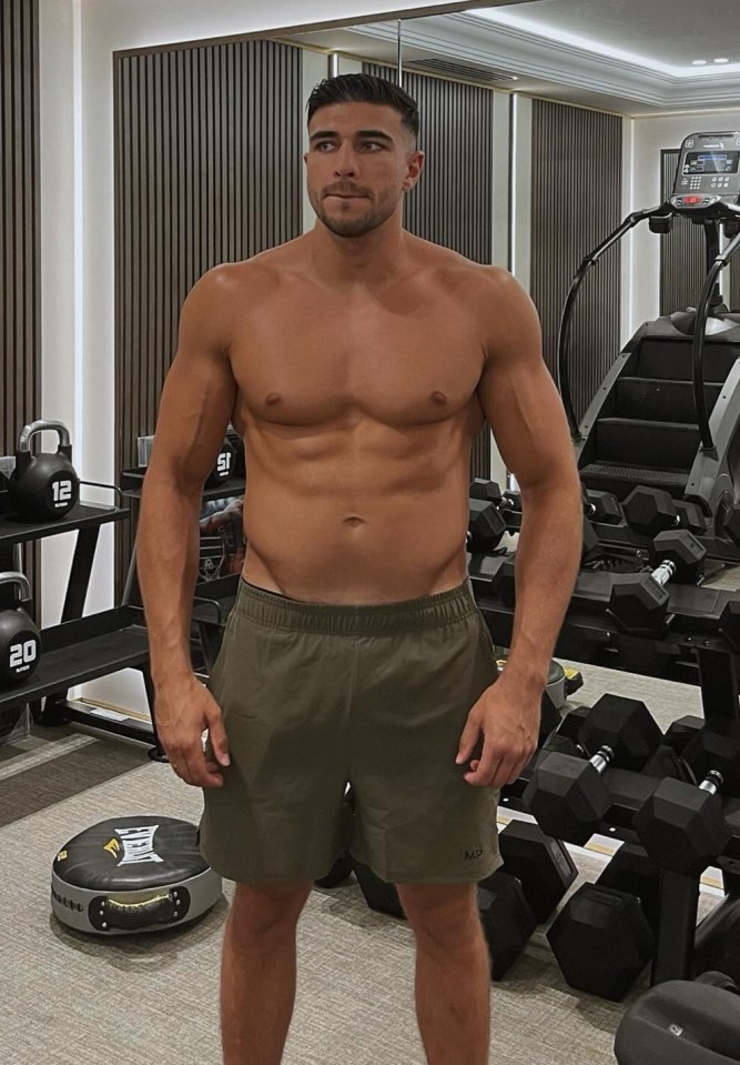 a shirtless man stands in a gym surrounded by dumbbells with the number 20 on them