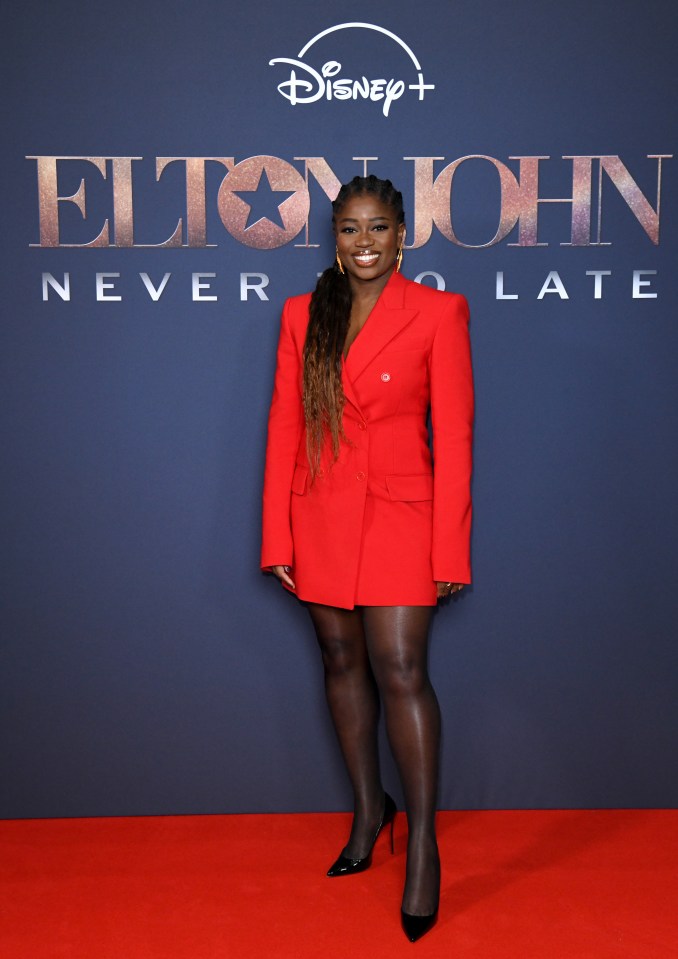 a woman in a red suit stands on a red carpet in front of a poster for elton john never too late