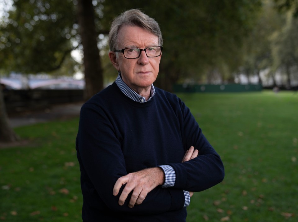 a man wearing glasses and a blue sweater stands with his arms crossed in a park