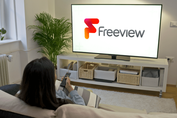 a woman sits on a couch watching a tv with the freeview logo on the screen