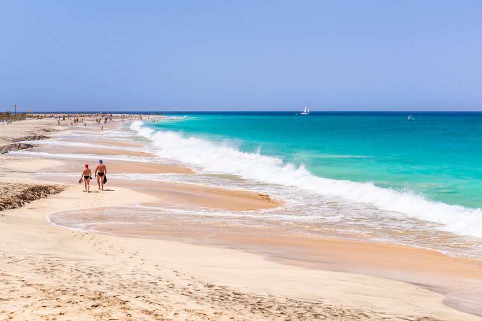 two people walking on a beach with a sailboat in the distance