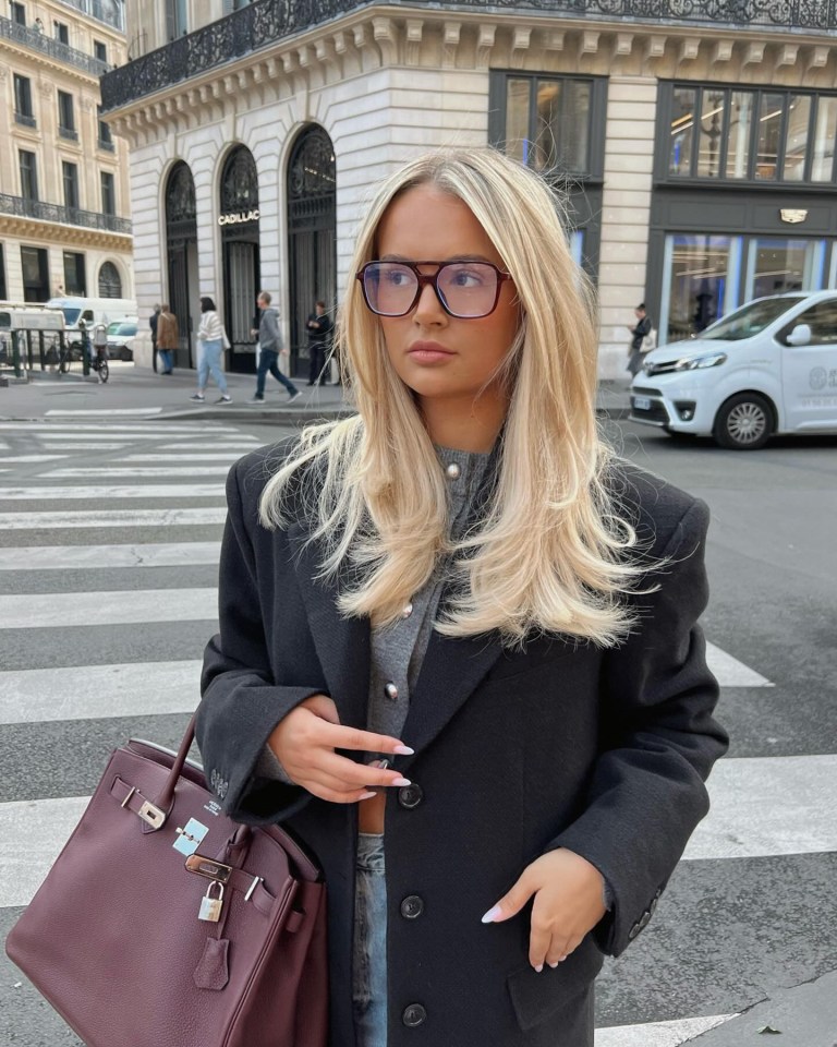 a woman wearing glasses and a black coat is crossing the street in front of a cadillac store