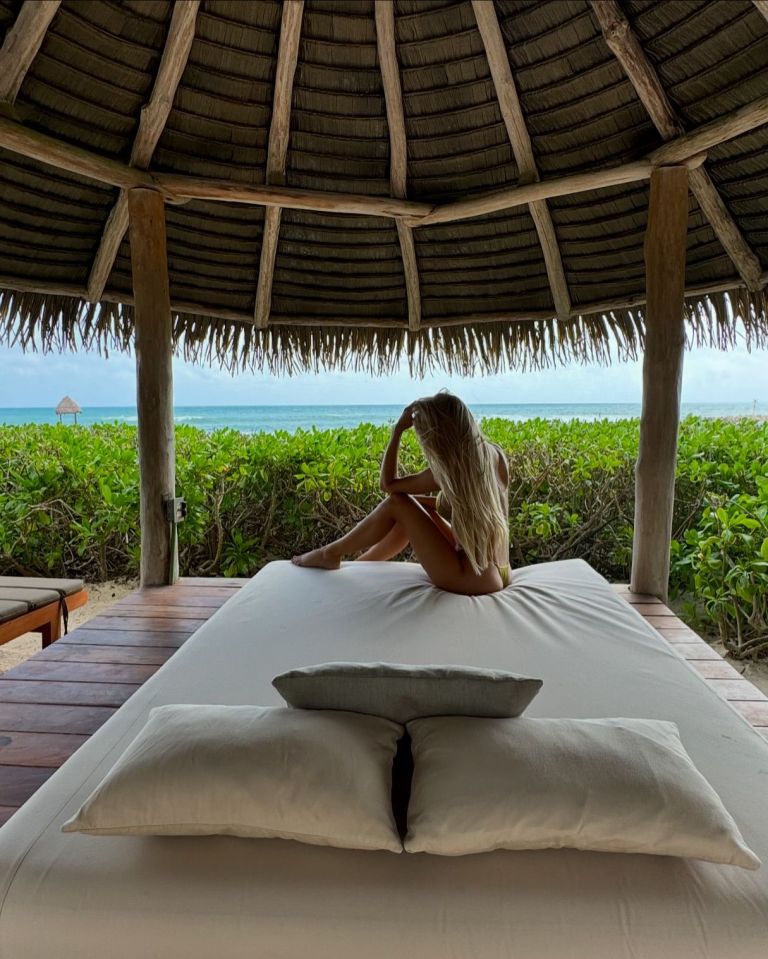 a woman sits on a bed under a thatched roof overlooking the ocean