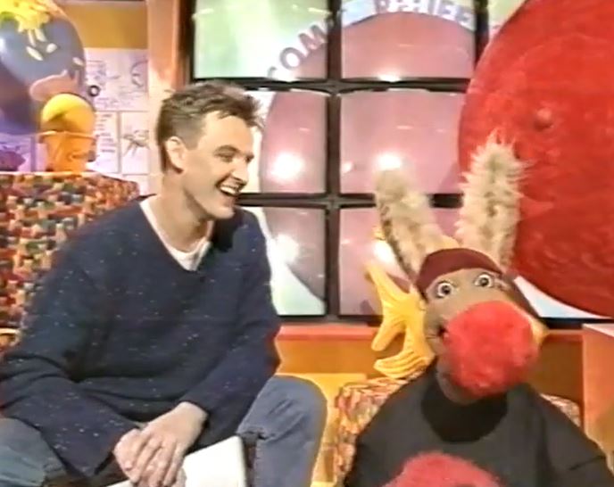 a man in a blue sweater sits next to a stuffed animal with a red nose