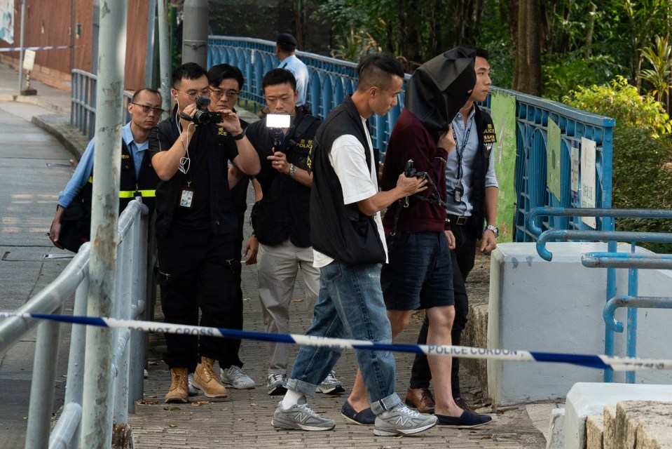 An unidentified person wearing a hood is escorted by police officers to a park for video reconstruction of a crime scene