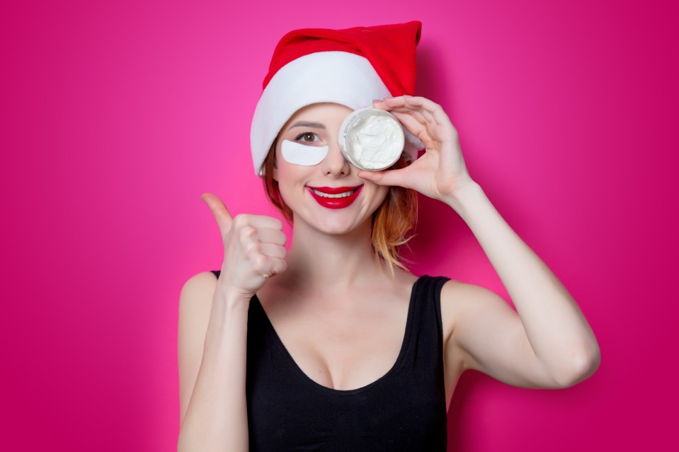 a woman wearing a santa hat is holding a jar of cream in front of her eye