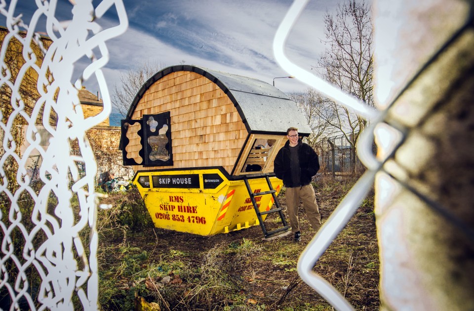 A man stands outside a small house built into a yellow skip.