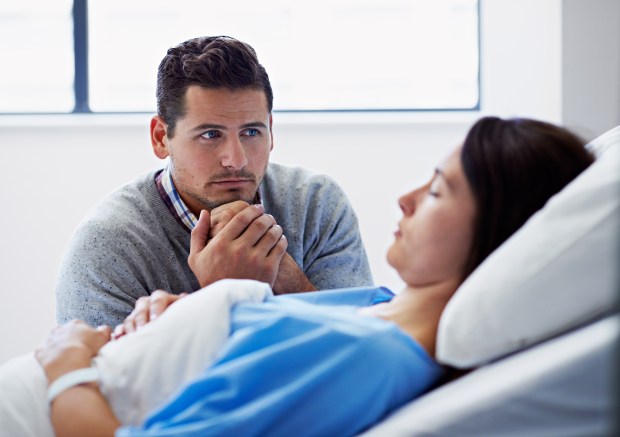 A man holds the hand of an unconscious woman in a hospital bed.