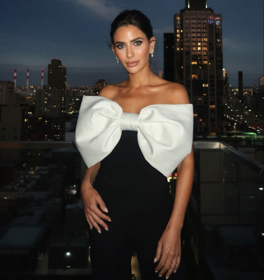 Woman in black jumpsuit with large white bow, posing on a rooftop at night.