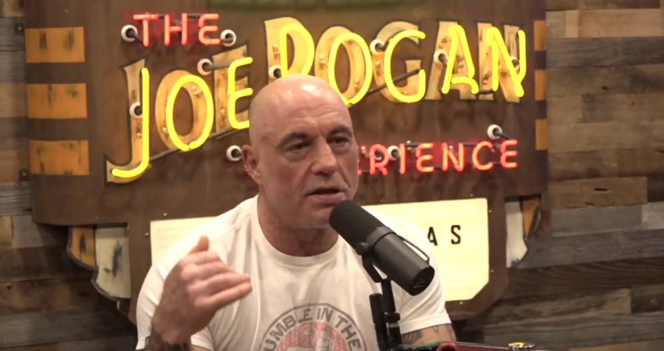 Joe Rogan speaking into a microphone in front of a neon sign for "The Joe Rogan Experience."