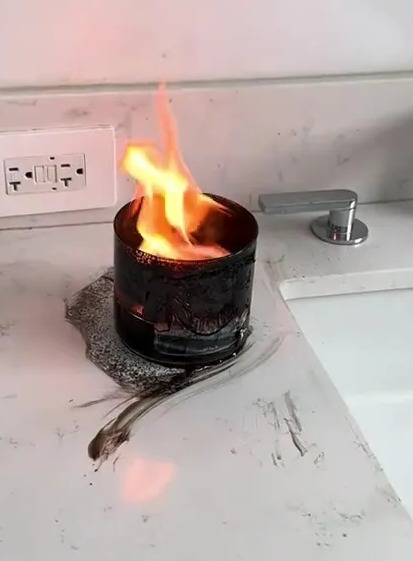 A woman is holding the remains of a shattered glass candle with wax and wick inside, with burn marks on the floor and walls behind her.