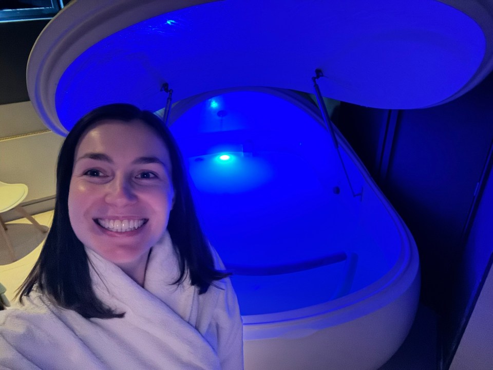 Woman in bathrobe smiling in front of a blue-lit floatation tank.