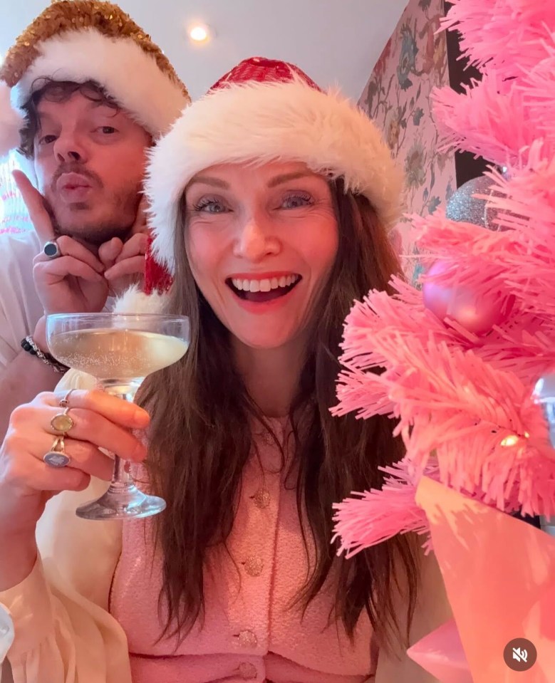 Couple in Santa hats toasting with champagne in front of a pink Christmas tree.