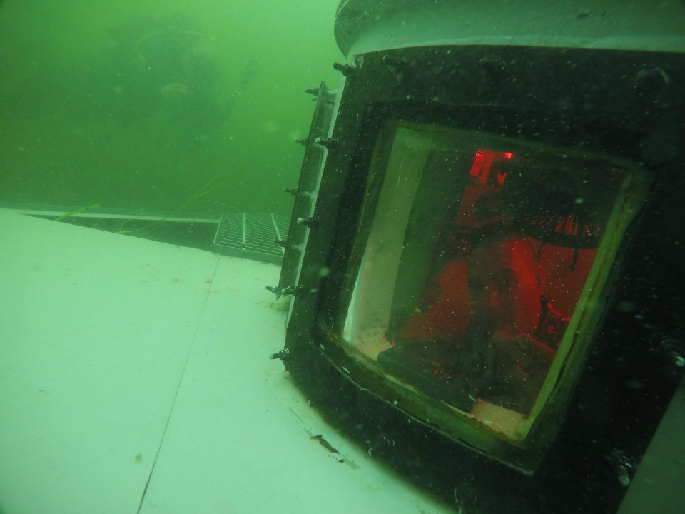 Underwater view of a submerged structure with a window showing a person inside.