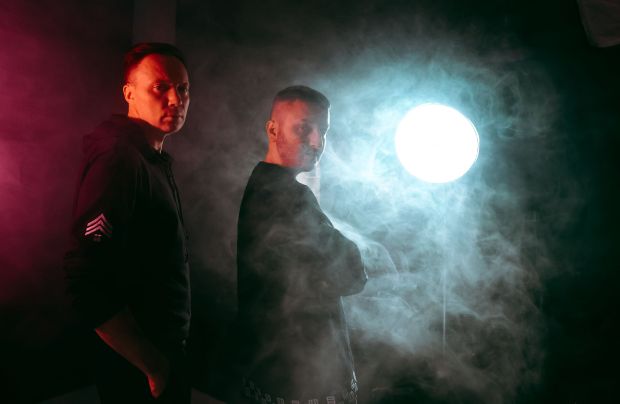 Two men in dark hoodies stand in smoky, dimly lit room.