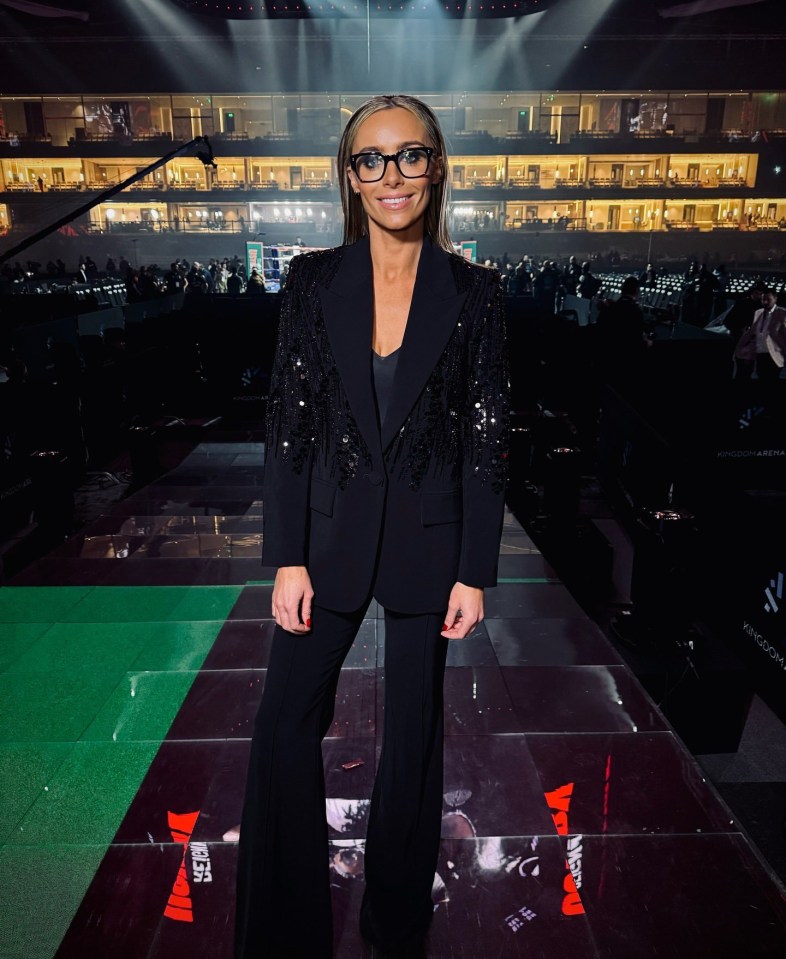 Woman in black suit and glasses standing in an arena.