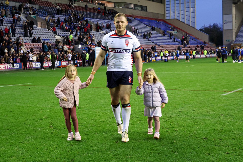 A rugby player walks off the field, holding hands with two young girls.