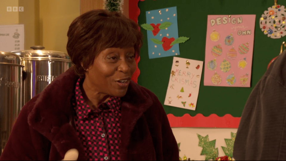 Woman in a burgundy coat smiling, standing in front of a notice board with children's Christmas artwork.