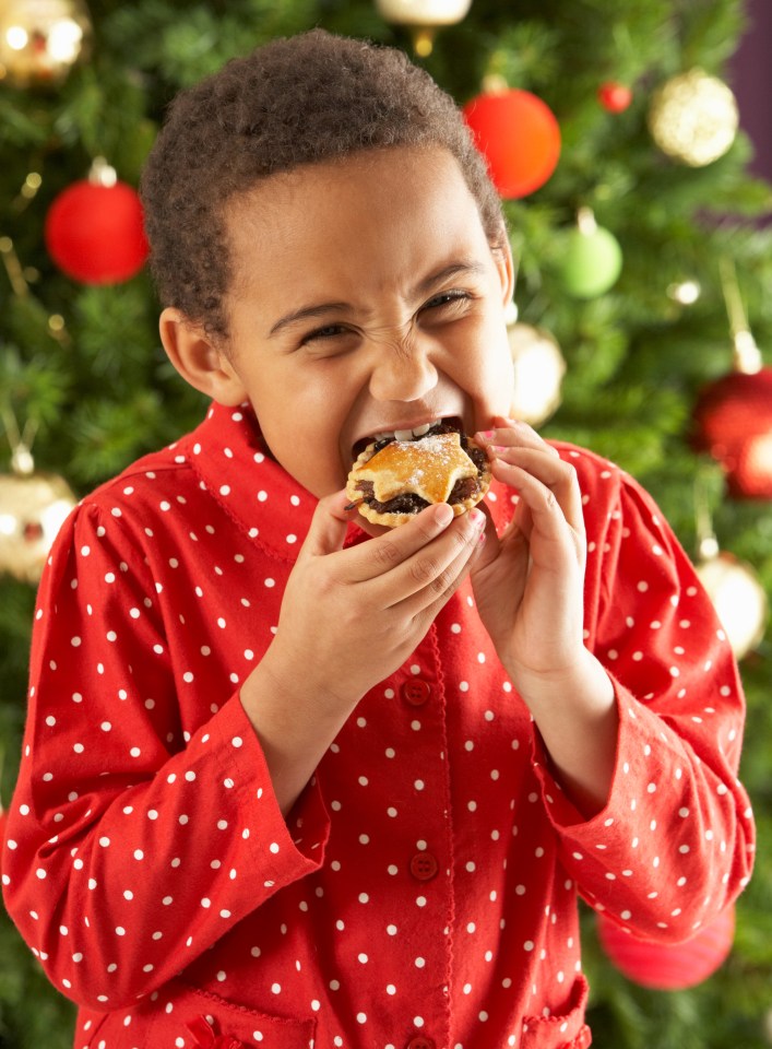 Child in red polka dot pajamas eagerly eating a mince pie in front of a Christmas tree.