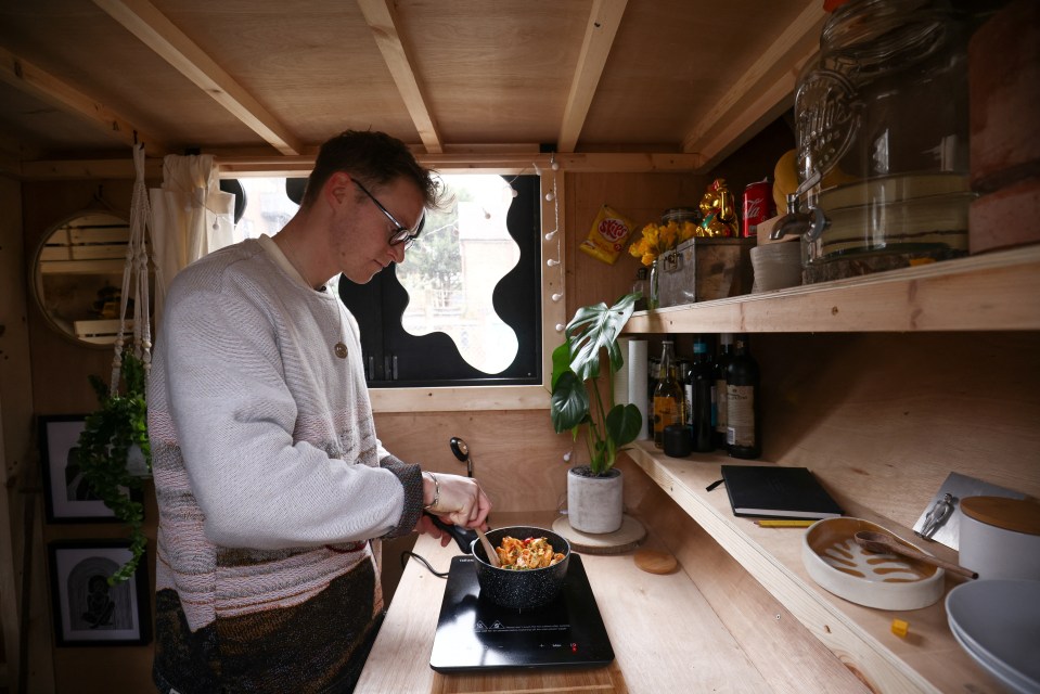 A man cooks a meal on a portable electric stove in a small, wooden room.