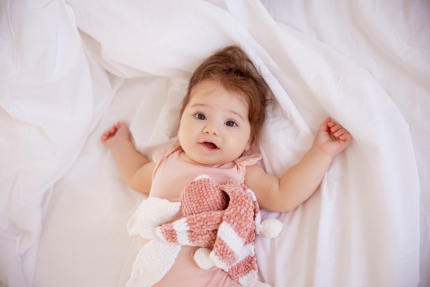 Baby girl in pink bodysuit lying on bed, holding a plush toy.