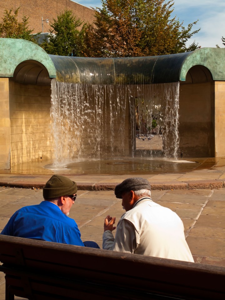 Two elderly men sitting on a bench, talking in front of a water feature.