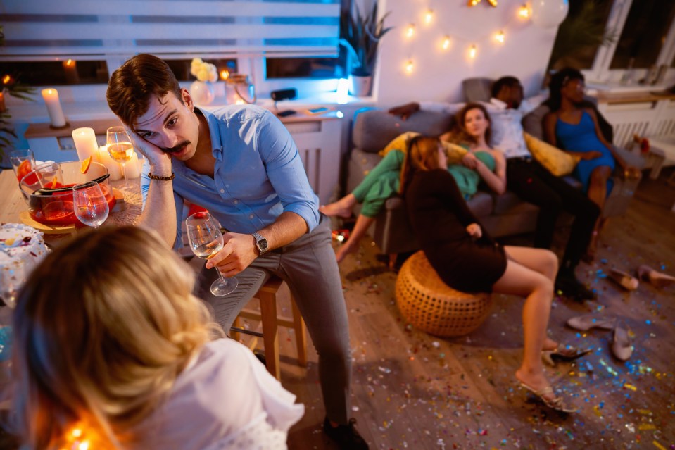 A man and woman flirting at a house party while other partygoers sleep on the furniture.