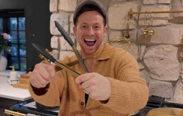 Man in a tan cardigan holding kitchen utensils in front of a range.