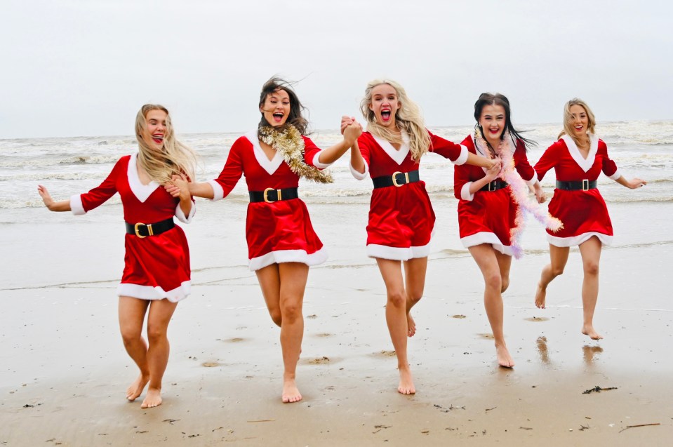 Five dancers in Santa Claus dresses run on a beach.