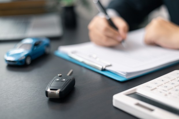 A car key and paperwork on a desk, with a person signing a document in the background.