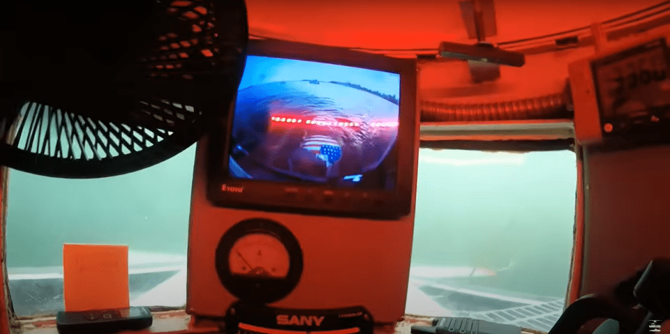 Interior view of a submersible showing a monitor displaying an underwater scene with an American flag.