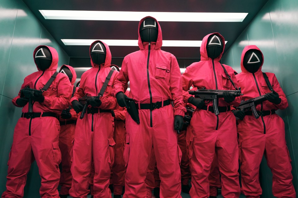 Squid Game guards in pink jumpsuits and masks holding guns in an elevator.