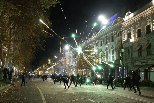 Protesters clashing with police amidst fireworks in a city street at night.