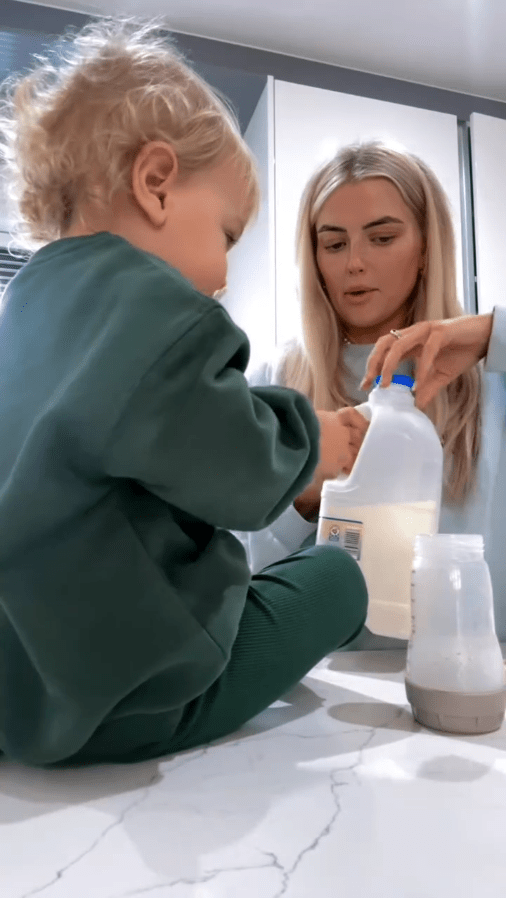 A toddler and a woman pour milk from a jug into a baby bottle.