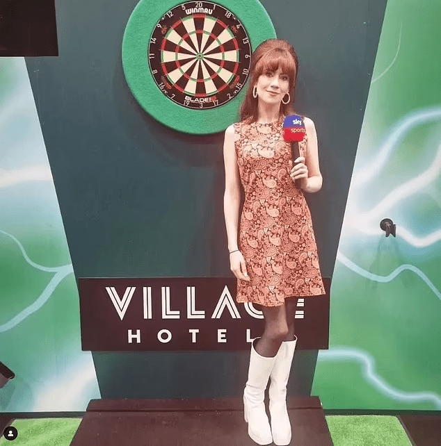 Woman in a 60s-style dress holding a microphone in front of a dartboard and Village Hotel sign.