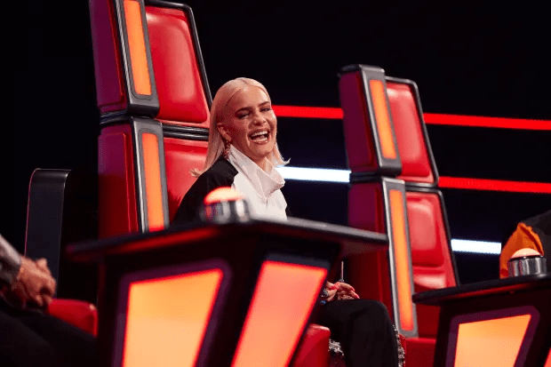 A smiling judge sits in a red spinning chair during a televised singing competition.