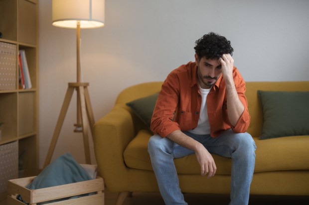 A worried young man sits on a sofa, hand on his face.