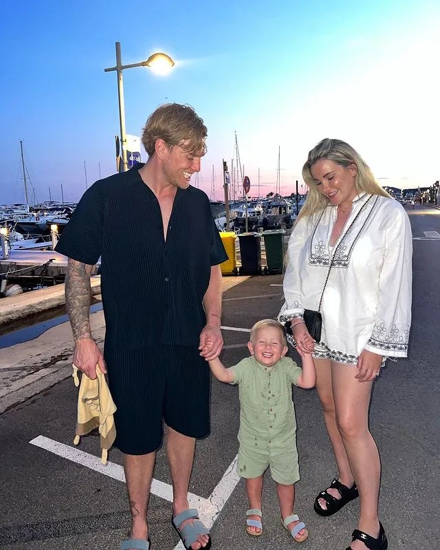 A family of three, a man, woman, and toddler, holding hands near a marina at sunset.