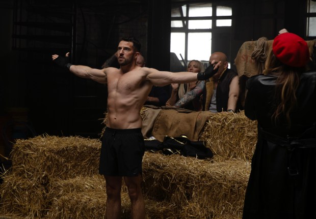 A muscular man in boxing shorts stands with arms outstretched in a makeshift boxing ring, watched by other people.