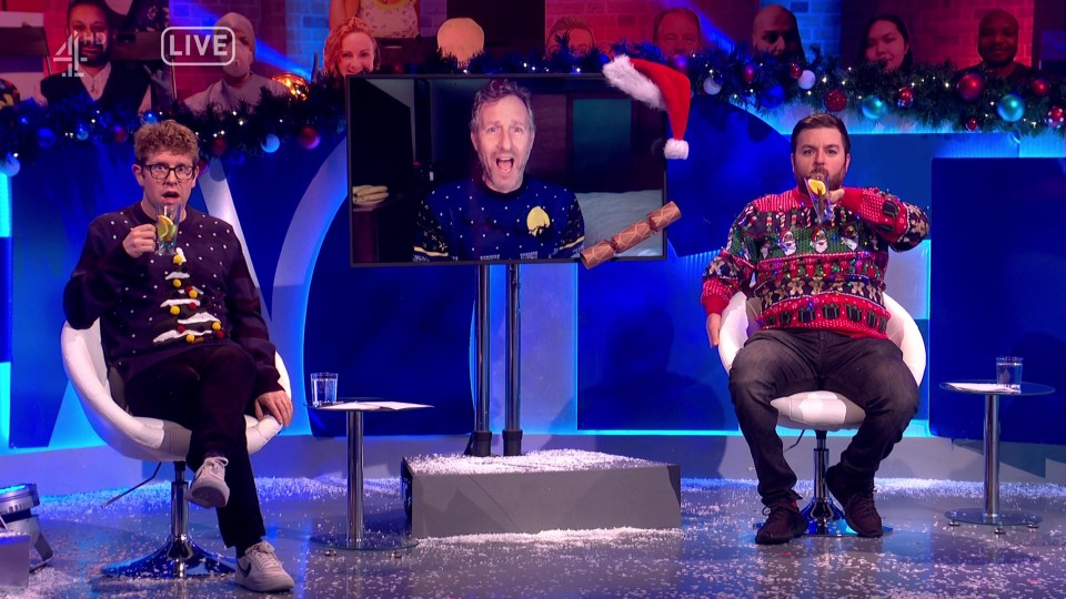 Two men in Christmas sweaters on a TV show, one drinking from a glass, with a guest appearing on a screen.