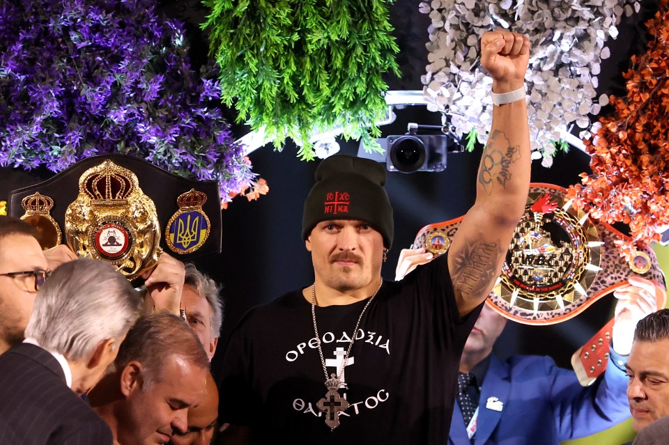 Oleksandr Usyk at a weigh-in, holding championship belts and raising his fist.