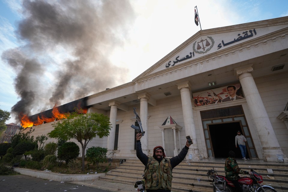 A man celebrates in front of a burning building, holding a rifle and a phone.