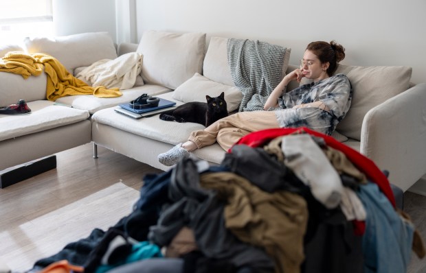 A woman relaxes on a couch with her black cat, while a pile of unfolded laundry sits nearby.