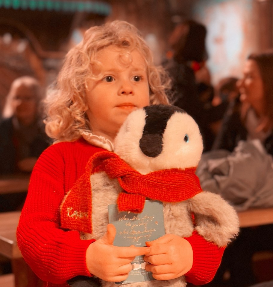 Child in red sweater holding a penguin plush toy and a book at Lapland UK.